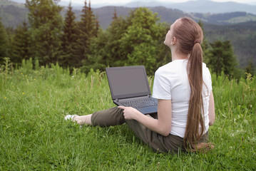 Beautiful dreaming girl with a laptop sitting on green grass on a background of mountains
