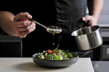 Close-up of the hands of a male chef on a black background. Pour sauce from the spoon on the salad dish.