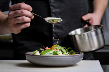 Close-up of the hands of a male chef on a black background. Pour sauce from the spoon on the salad...