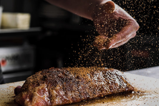 Raw piece of meat, beef ribs. The hand of a male chef puts salt and spices on a dark background.