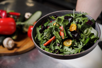 Cook man in a black apron holds a dish of salad in his hand. Against the background of the kitchen of the restaurant.