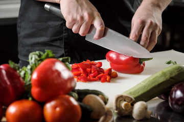 Male chef cuts vegetables for salad in a restaurant in a black apron. White cutting board, closeup of hands.