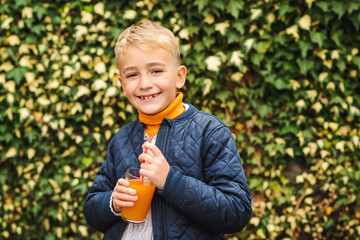 Happy cute boy drinking juice. Kid holding glass of orange juice at greenery background. Fresh...