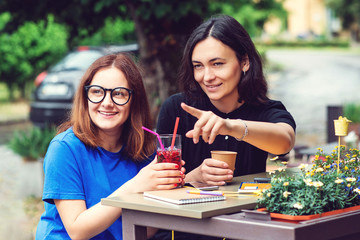 Two excited young girls talking in summer cafe outdoors. Happy friends. Young women drinking juice and enjoying in conversation. People, consumerism, lifestyle concept.