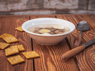 Lunch from a broth with slices of boiled turkey, quail eggs in a deep white plate are on a wooden table. A knife and spoon, a few crackers with herbs lie near the plate.