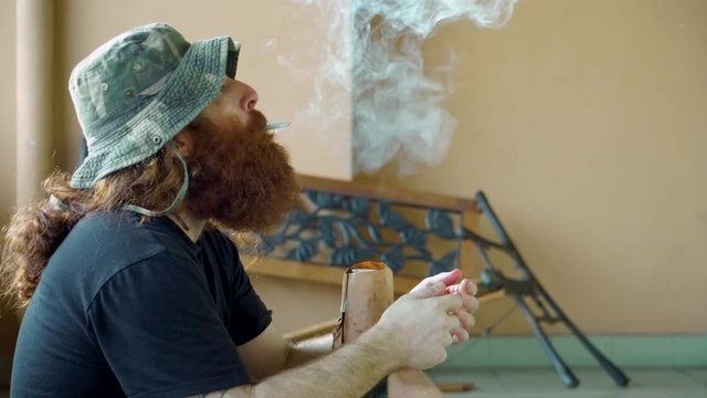 Homeless Man lighting up marijuana or tobacco cigarette and drinking beer can outdoors near broken bench