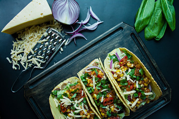 Photo of mexican tacos with pork meat, cheese, corn, onions and herbs on a wooden board. Sauce ketchup and hot pepper. Black background. Side view.