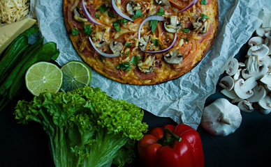 Fresh pizza with sausage, tomatoes, mushrooms and cheese on a dark background. Decorated with fresh vegetables and spices. View from above.