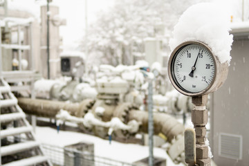 Closeup of manometer high pressure meter measuring pressure of natural gas on industrial site with valves and pipes on the background. The meter is covered with snow in cold winter. Selective focus.