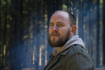 Portrait of contented Adult young man in a tourist suit on nature in the spring forest on the background of tree trunks and blue smoke from a fire