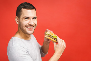 Young man holding a piece of hamburger. Student eats fast food. Burger is not helpful food. Very hungry guy. Diet concept. Isolated over red background.