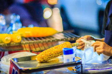 Close up hand of street food vendor while grilling and selling for mixed sweet corn butter. Cook is selling grilled sweet corn on street in Bangkok night market, Thailand.