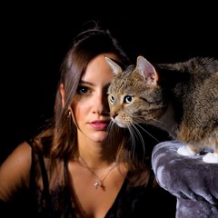 A young girl poses with her pet cat in front of a black background. She poses in the background and puts  her cat prominent in the foreground.