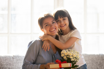Happy kid daughter embracing mom holding flowers bouquet and gift