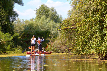 Athletic teen girl and man paddling on the SUP (stand up paddle board, paddleboard) in wilderness river overgrown duckweed near thickets of trees and wildgrapes at summer