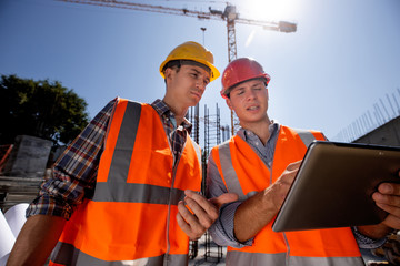 Structural engineer and construction manager in orange work vests and hard helmets discuss...