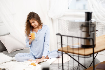 Beautiful brunette girl dressed in the light-blue pajama sits on the bed with glass of fresh juice...