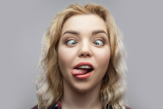 Portrait of funny crazy young beautiful blonde woman in casual red checkered shirt standing and looking with crossed eyes and tongue out. indoor studio shot, isolated on grey background.