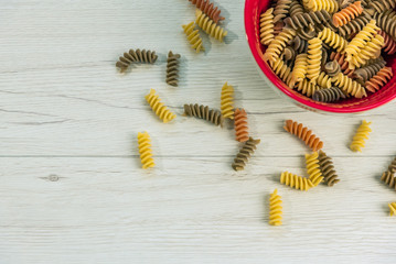 Naklejka premium Raw uncooked three-colored Fusilli in a red bowl on white wood table.