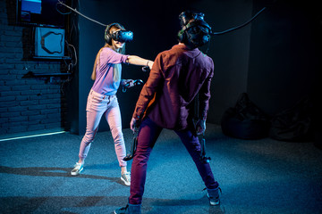 Fototapeta premium Man and woman playing game using virtual reality headset and gamepads in the dark room of the playing club