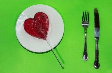 Red heart lollipop ready to eat on a white plate with stainless steel fork and knife
