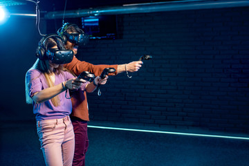 Man and woman playing game using virtual reality headset and gamepads in the dark room of the...