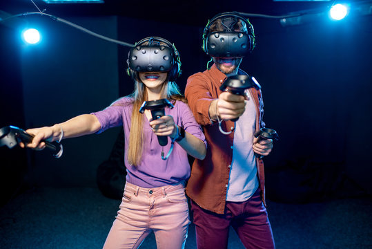 Man and woman playing game using virtual reality headset and gamepads in the dark room of the playing club