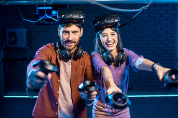 Portrait of a young couple standing together with virtual reality headsets and gamepads ready to...