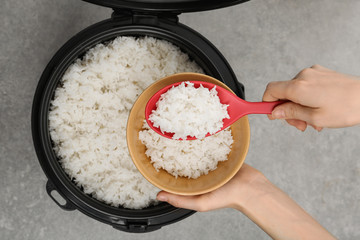 Woman putting boiled rice into bowl from cooker on grey background, top view