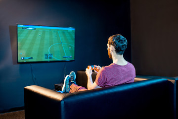 Man playing football game with gaming console sitting on the couch in front of the monitor at home...