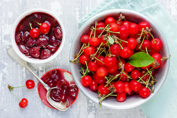 Homemade cherry jam in sugar syrup in glass jar on table with fresh cherries in bowl top view