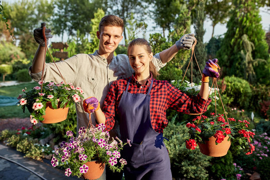 Happy guy and girl gardeners hold pots with petunia in the wonderful garden on a sunny day.
