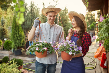 Happy guy and girl gardeners in a straw hats hold pots with petunia on the garden path in  on a sunny day.