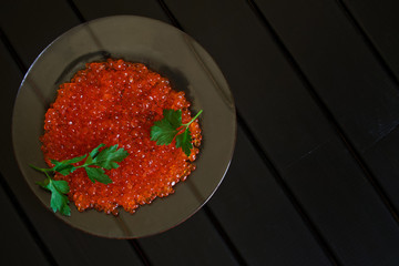 Red caviar of salmon fish and parsley leaves in a transparent plate on a black background