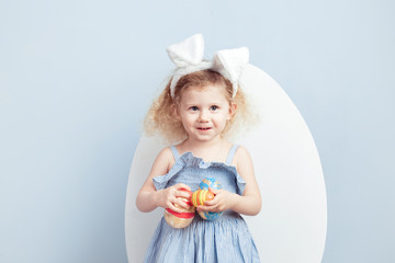 Charming curly girl in the light-blue dress with bunny ears on her head holds the dyed eggs in her hands on the background of a big white egg on a blue wall. Easter bunny