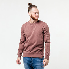 Fototapeta premium Fashionable man with beard and bun hairstyle dressed in light-brown long sleeve sweater and jeans poses in the studio on the white background