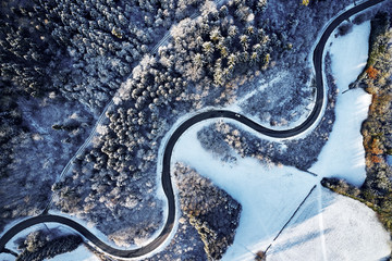 Aerial drone view of a curved winding road through the forest up in the mountains in the winter with snow covered trees and curved streets in winter while sunset and sun rays casting trough