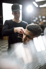 The fashion barber in black clothes makes a razor cut hair for a black-haired man sitting in the...
