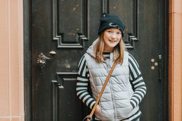 Outdoor portrait of young kid girl wearing grey sleeveless down jacket and black hat with cat ears