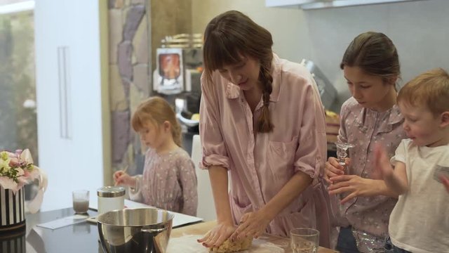 Mother takes huge piece of dough and tries to show her kids how to knead a dough. They are excited to learn and help. Eldest daughter brings a glass for making circles and baking them into cookies.