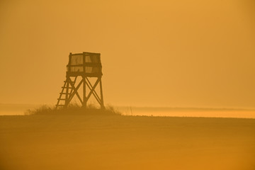 A silhouette of a watchtower in a landscape in the morning sunset. The whole pictures is in a golden orange colour.