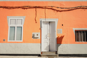 Fototapeta na wymiar A facade of a vibrant orange house with a window and an open white front door in Galdar, Gran Canaria