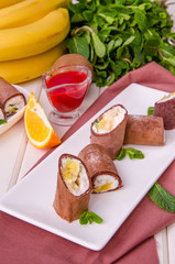 Sweet chocolate rolls from pancakes with cream cheese, fresh fruit and berry sauce on a white wooden background.