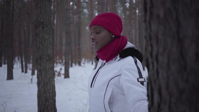 African american girl dressed warm wearing a red hat, scarf and white jacket standing in winter forest looking in side with her hand on the tree then turns and looks back. Slow motion shooting