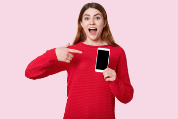 Image of European positive young woman points at cell phone with empty screen, dressed in red sweater, advertises new gadget, poses over light rosy background. Technology and people concept.