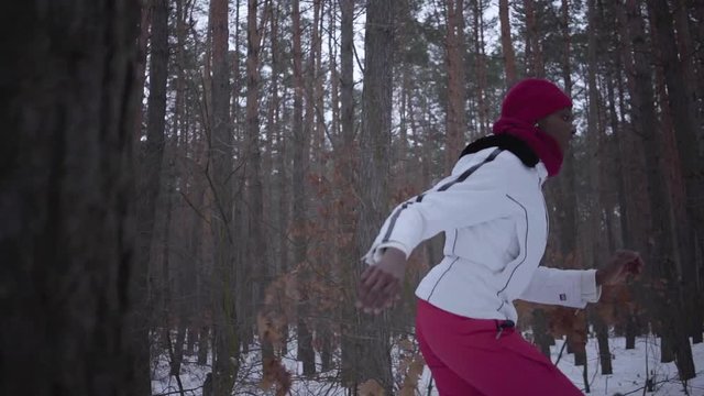 Scared African american young woman dressed warm wearing a red hat, scarf and white jacket running through the snowy forest. The girl constantly looks back running away from the pursuer. Slow motion.