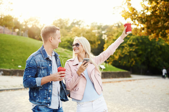 Smiling Couple In Love Walking In Autumn Park, Holding Hands. Listen To Music On Headphones And Drink Coffee To Go. Have A Fun Together. Hugging And Kissing 