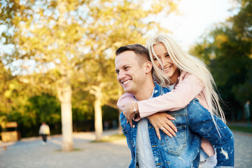 Fototapeta premium Happy couple on vacation. Lovers are laughing. Happy guy and girl. Lovers enjoy each other in the sunny park. Boyfriend carrying his girlfriend on piggyback