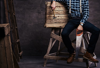 Worker, carpenter, handyman man wearing a checkered shirt with suspenders holding a hammer and saw...