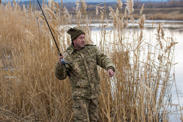 Fishing in the reeds for spinning on quiet water. A man in camouflage clothes in cool weather is fishing on the river bank.	